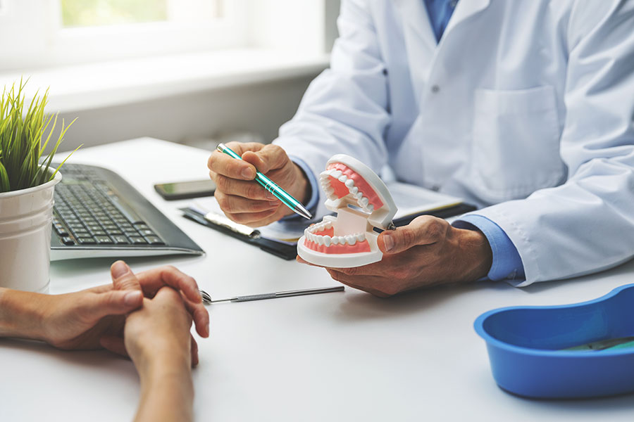 The image shows a dental professional, likely a dentist or orthodontist, examining a patient s teeth with a pair of gloves on and an open mouth model. They are seated at a desk in what appears to be a dental office setting, surrounded by typical dental equipment and tools.