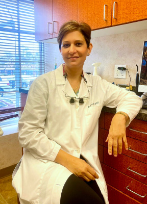 A woman wearing a white lab coat is seated at a dental office counter, posing for the camera with her hands on her knees.