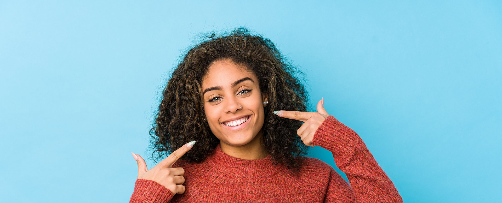 The image shows a person with a joyful expression, making a peace sign gesture with their right hand. They are smiling and appear to be indoors against a blue background.