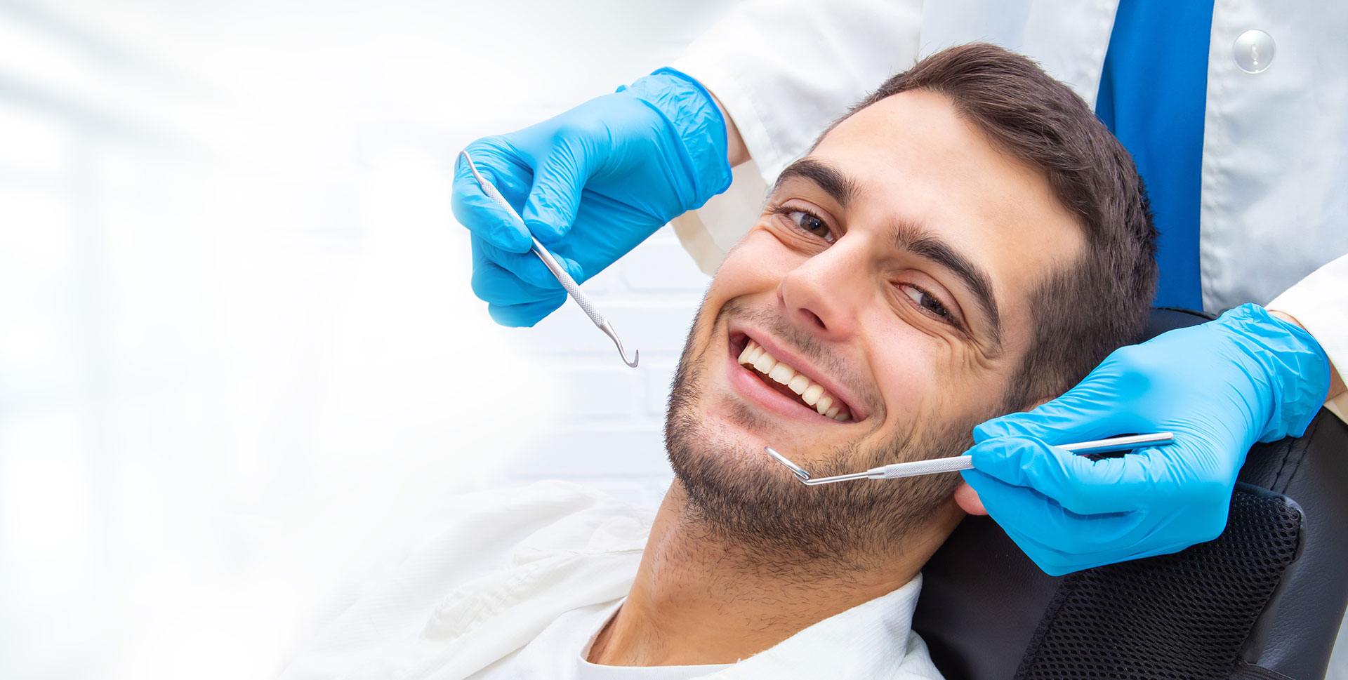 The image shows a smiling man in a dental chair, receiving dental care from a professional with a blue apron and gloves.