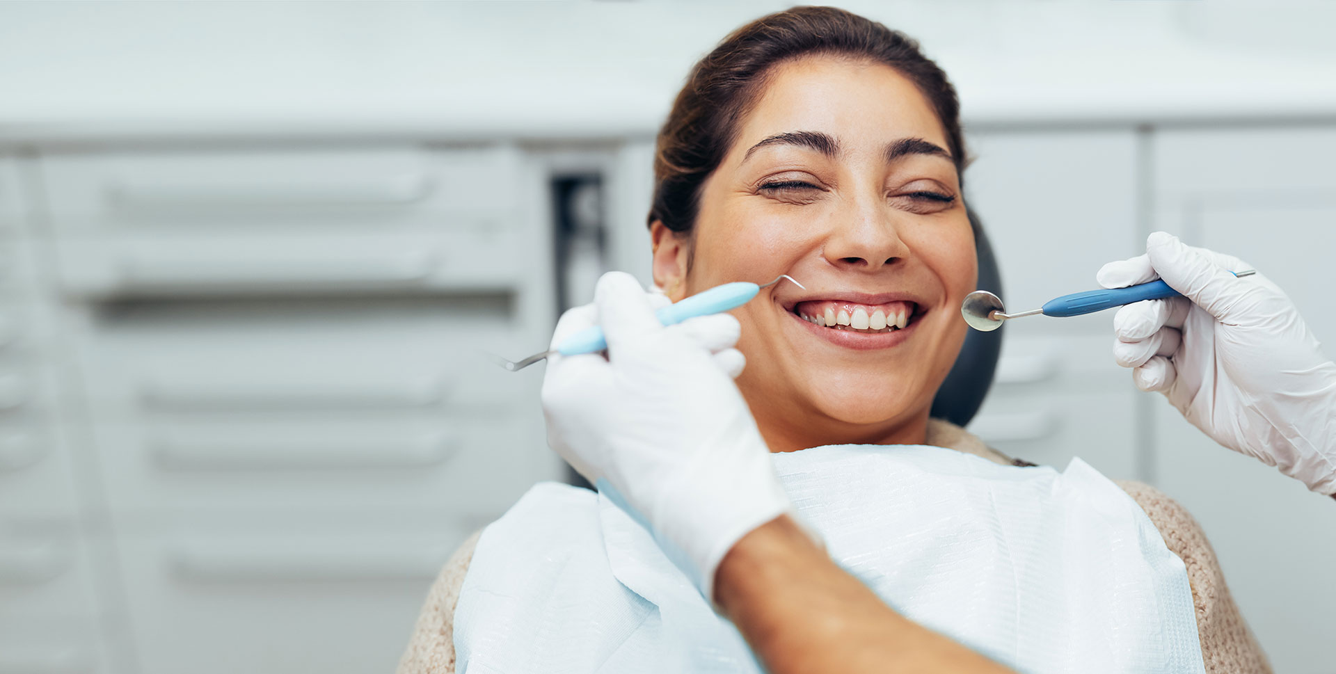 An image of a smiling woman sitting in a dental chair, receiving dental treatment from a dentist who is holding a toothbrush.