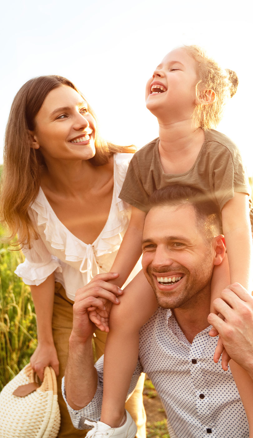 A family of three, with a woman and child smiling, and a man laughing, all enjoying an outdoor setting during sunset.