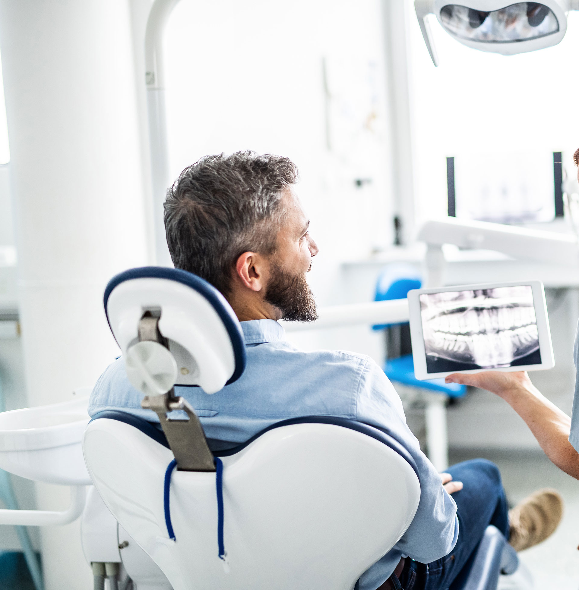 A man in a dental chair is being shown his teeth on a digital display by a dentist, with the man s reflection visible on a tablet.