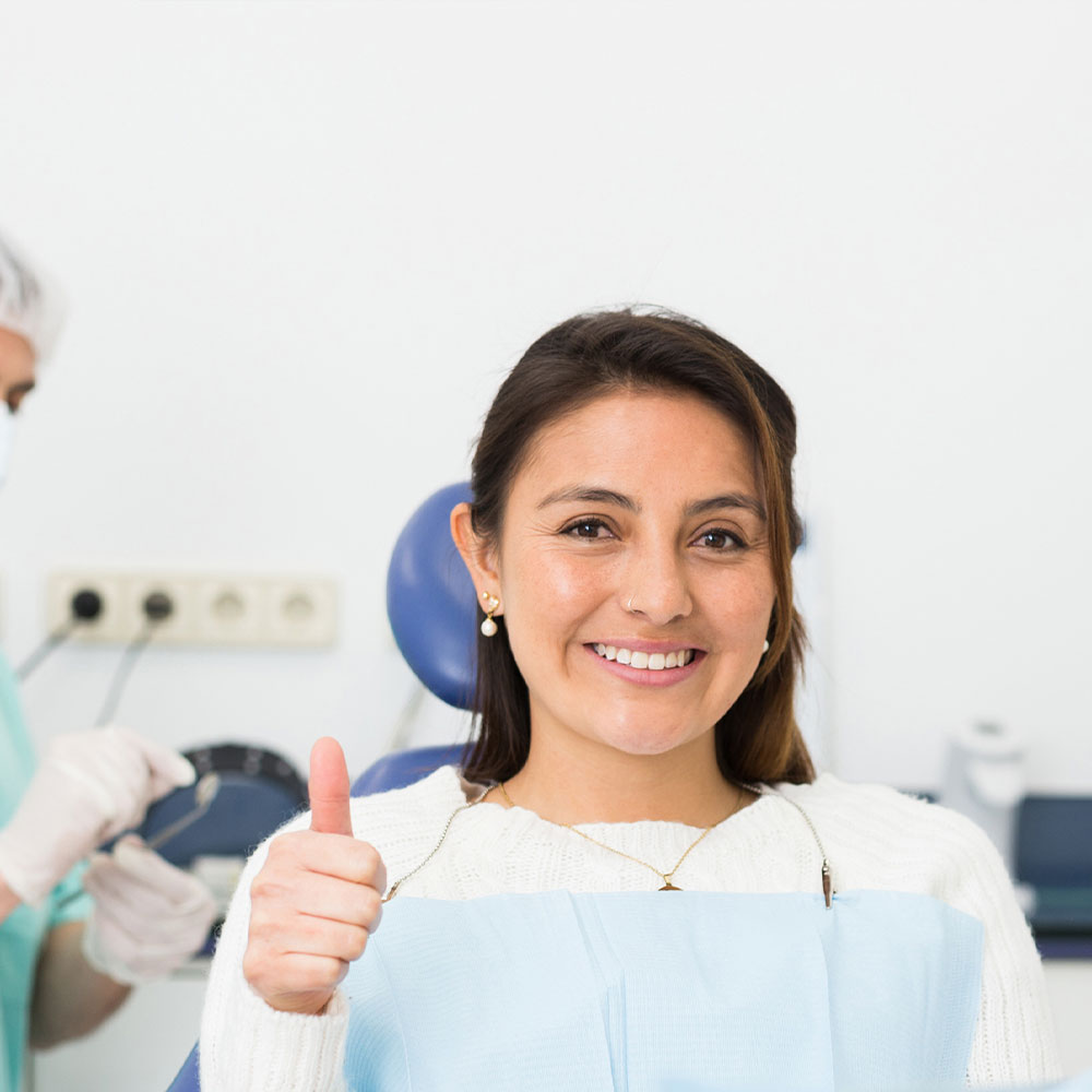 A woman in a dental office, smiling and giving a thumbs-up, with a dental hygienist attending to her.