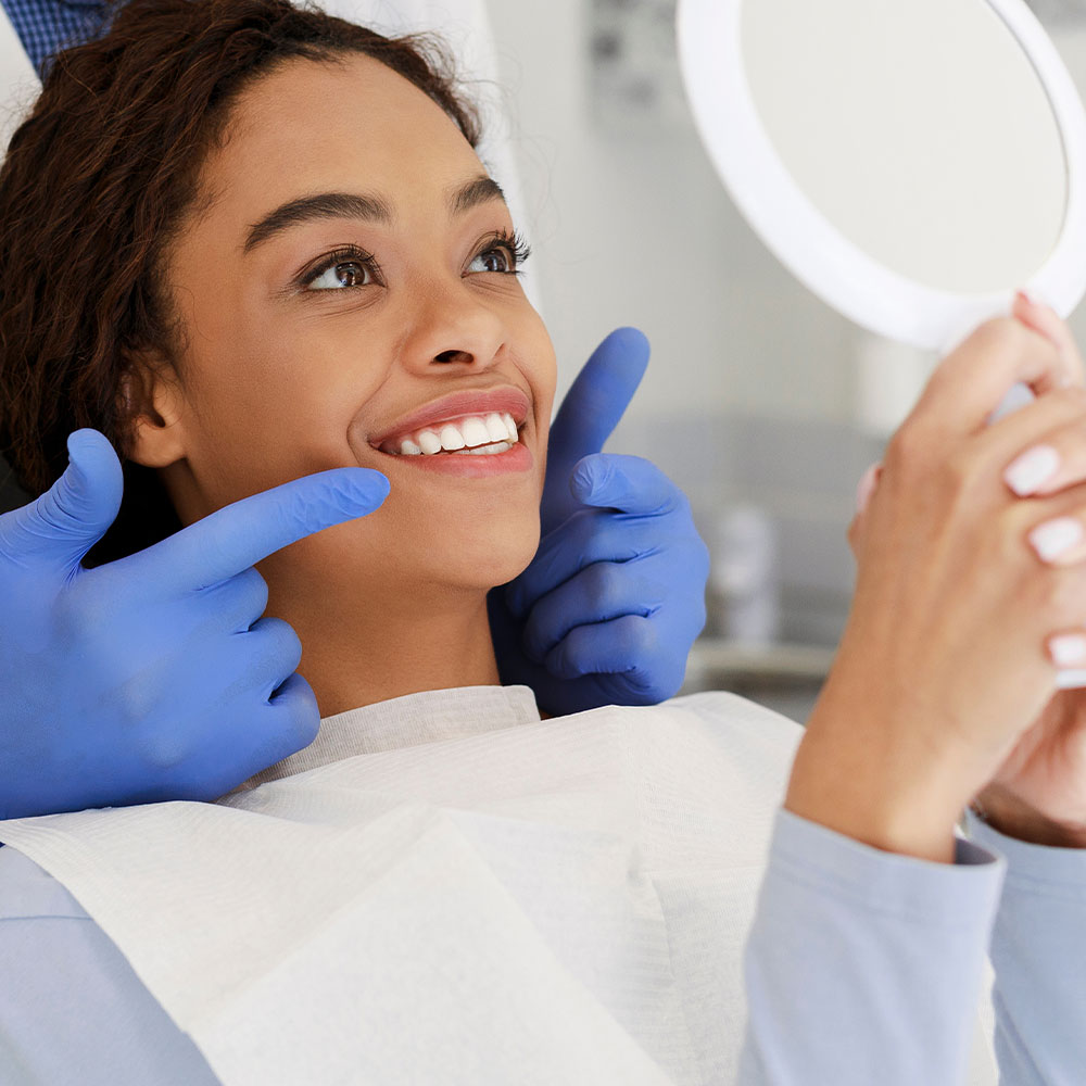 The image shows a young woman sitting in a dental chair, smiling at the camera with her eyes closed. She is wearing a blue surgical gown and has a mirror held up to her face by a dental professional, who appears to be adjusting or examining her teeth.