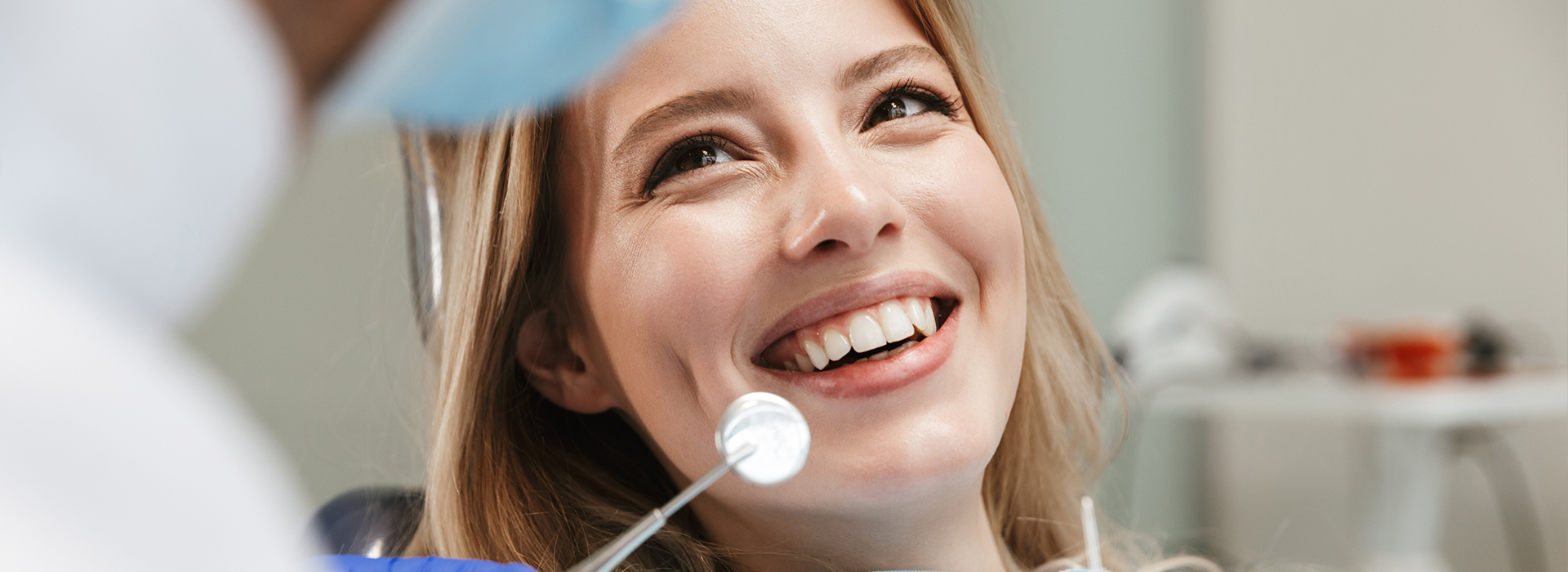 The image shows a woman smiling at the camera while seated in a dental chair, with a dentist standing behind her, both within a dental office setting.