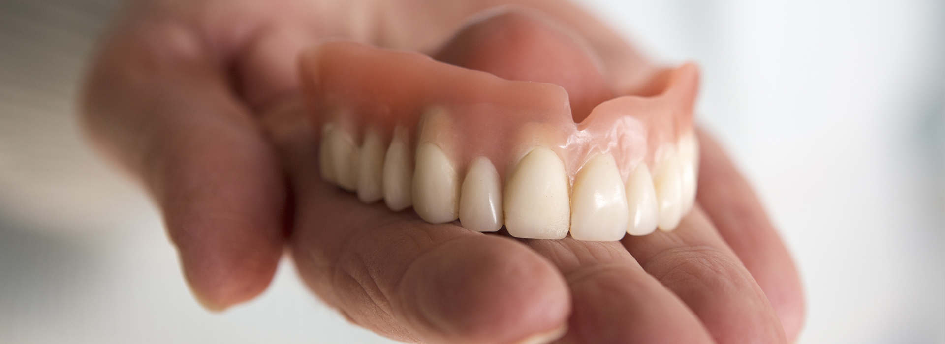 An adult human hand holding a set of artificial teeth with visible roots and gumline, showcasing dental prosthetics.