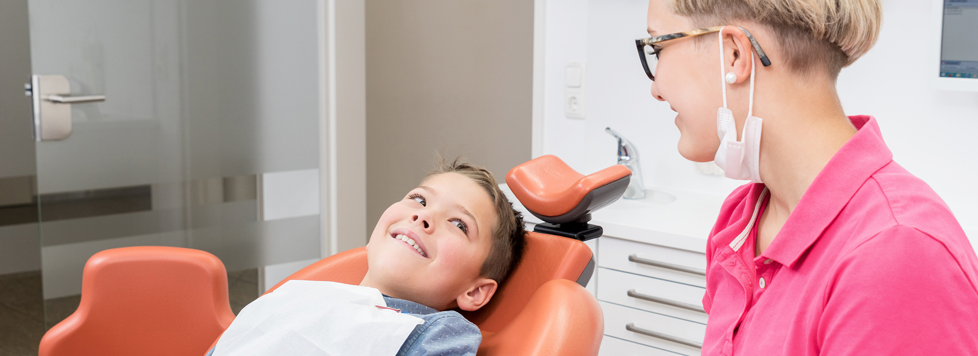 A woman and a child in a dental office, with the woman sitting at a desk and the child lying on a dental chair.