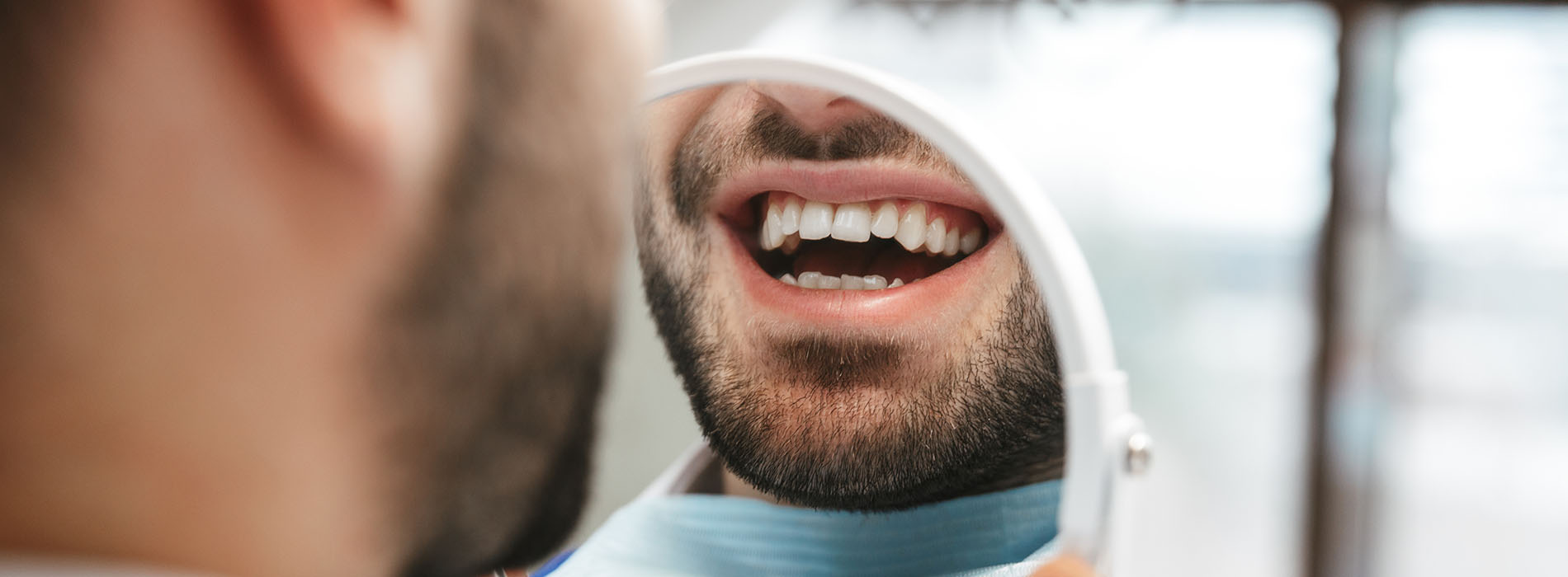 The image shows a man with a beard sitting in front of a dental mirror, smiling while looking at his reflection.