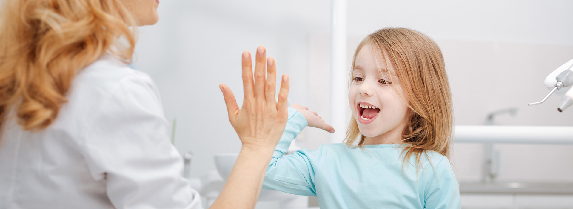 A woman and a young girl in a dental office, the woman gesturing with her hand while standing behind the child who is seated at a dental chair.