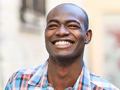 The image features a smiling man with short hair, wearing a light blue plaid shirt. He is standing outdoors in front of a building with a red and white striped awning.