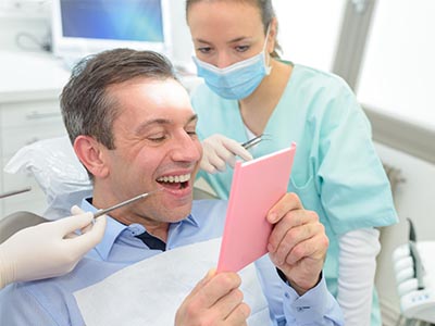 The image shows a man sitting in a dental chair, holding a tablet with a smile on his face, while a dentist and hygienist are attending to him.