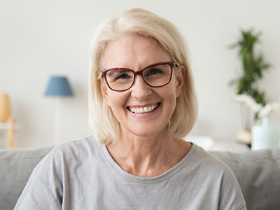 A smiling woman with short blonde hair, wearing glasses and a white shirt, sitting on a couch.