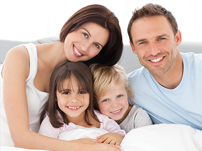 A family of four, including two adults and two children, posing together in a bedroom with a white bedspread.